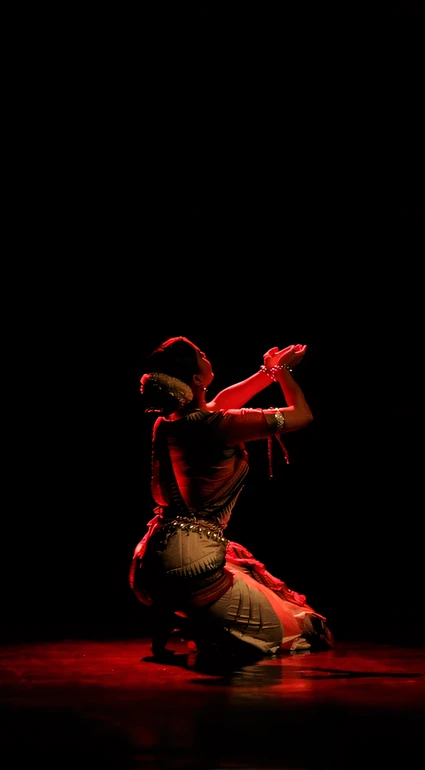 Surupa kneeling, bathed in red light, palms joined and raised upward as if receiving a boon